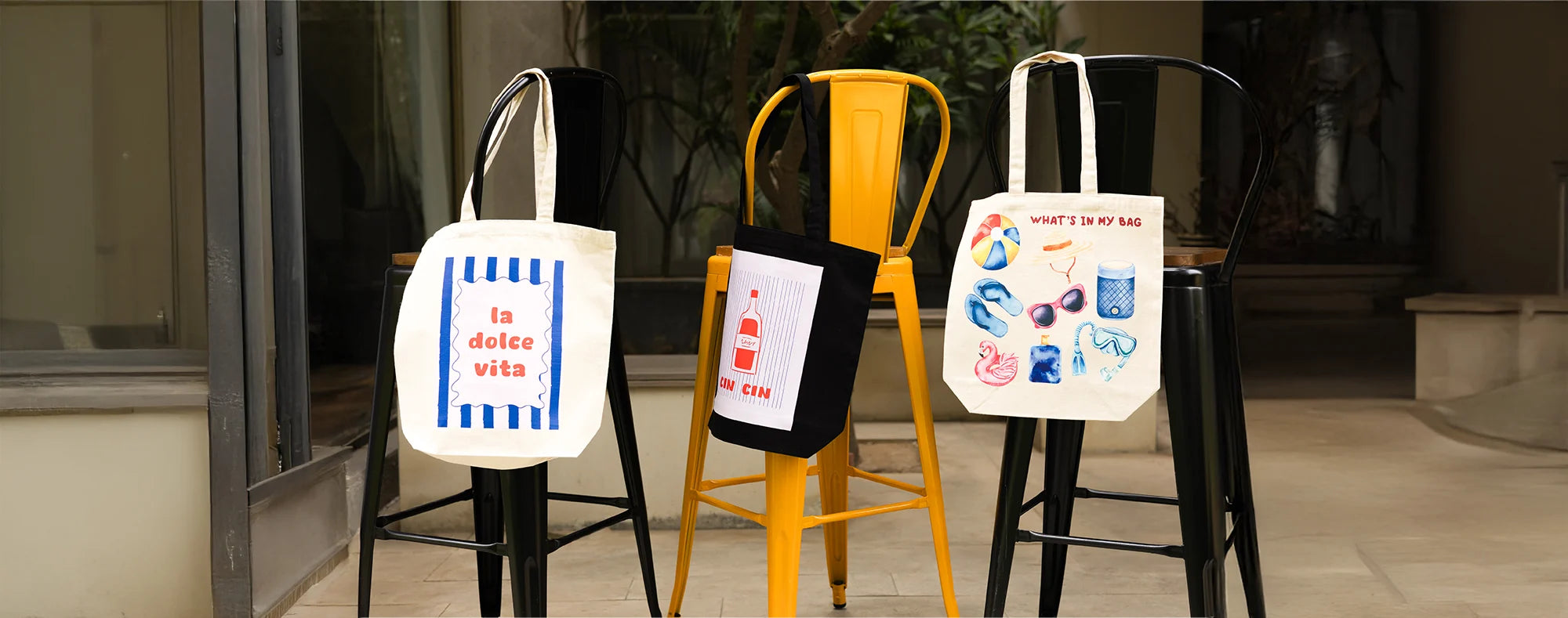 Four tote bags with different designs on black, yellow, and gray stools.